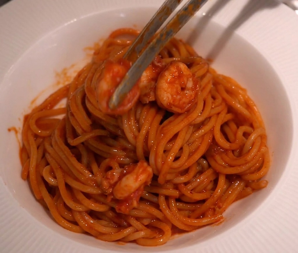 Pinzas de cocina colocando camarones sobre el nido de espaguetis en un bol blanco.
