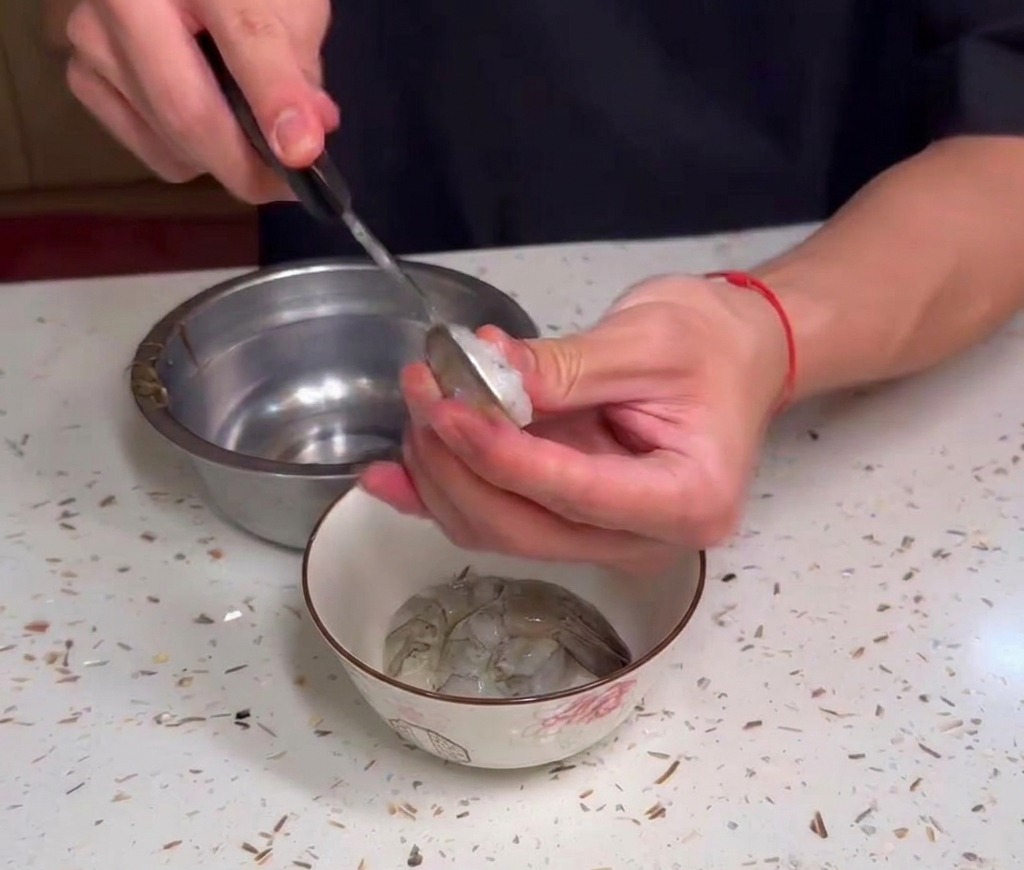 Manos limpiando camarones crudos sobre un pequeño bol en una encimera moteada.