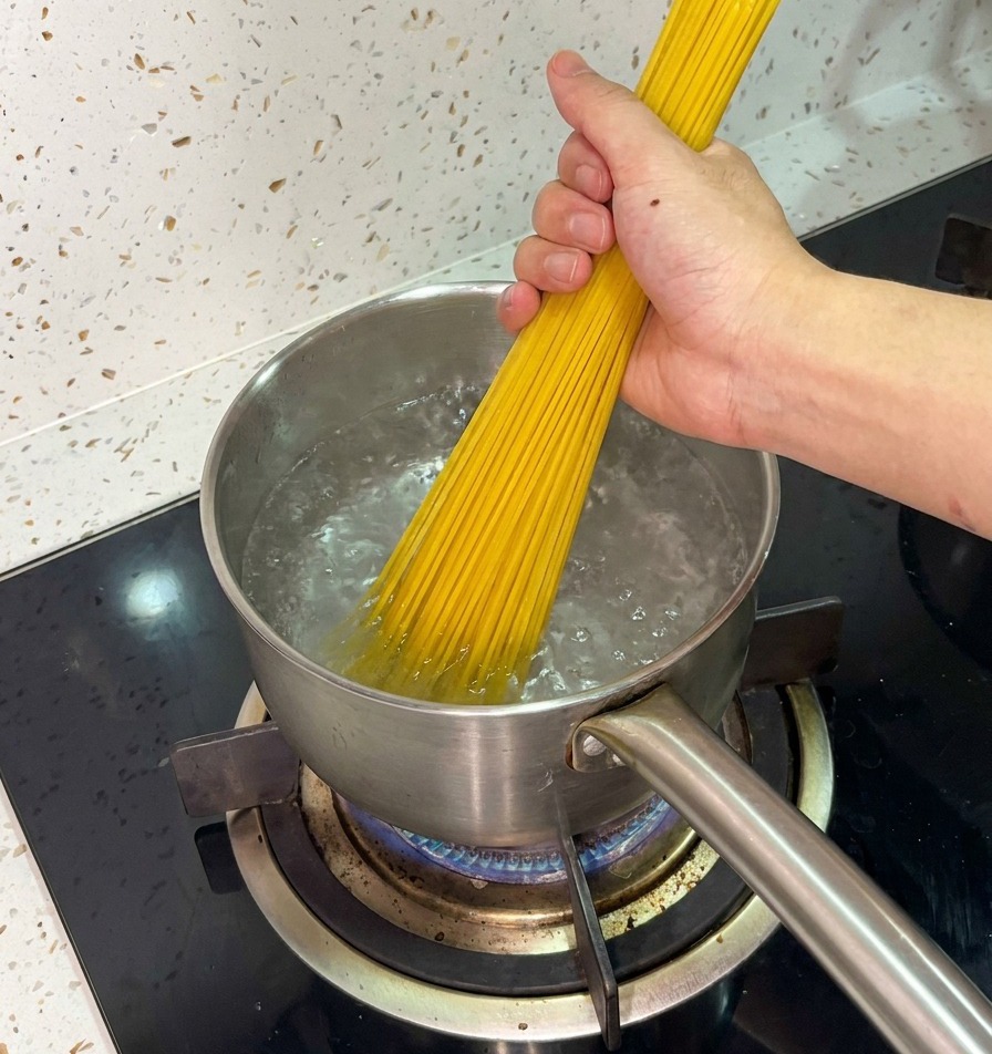 Una mano sujetando firmemente un manojo de espaguetis secos y colocándolos en una olla de acero con agua hirviendo.