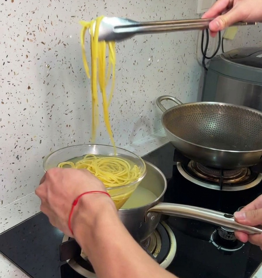 Pinzas de metal levantando espaguetis cocidos de una olla caliente y transfiriéndolos a un bol de vidrio con agua fría.