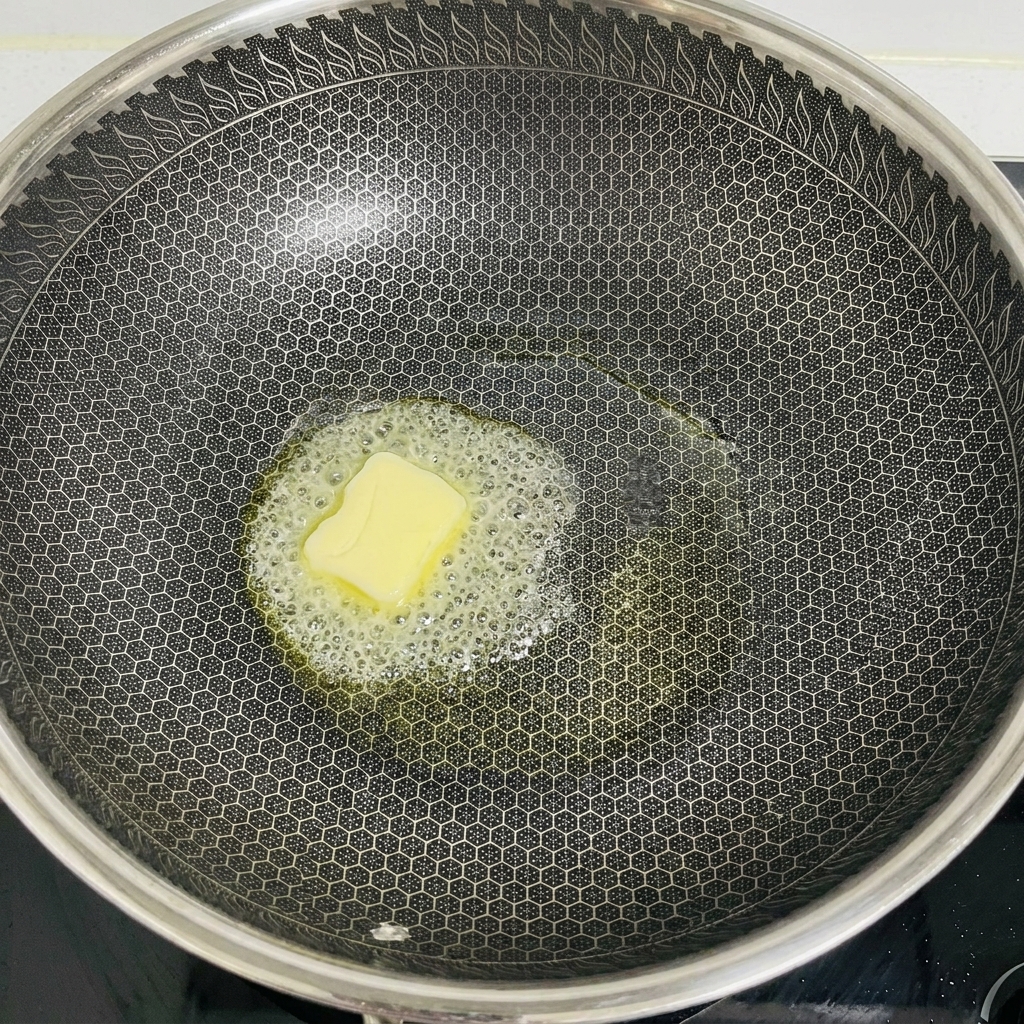 Un trozo cuadrado de mantequilla amarilla derritiéndose y burbujeando en el centro de un wok oscuro.