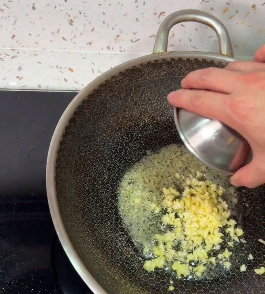 Una mano inclinando un pequeño bol de metal para dejar caer ajo recién picado en la mantequilla derretida dentro de un wok caliente.