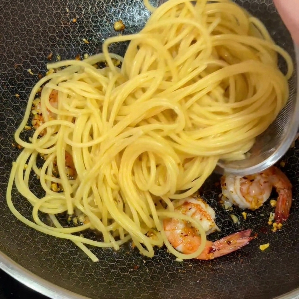 Fideos de espagueti cocidos siendo añadidos a un wok con camarones rosados y ajo picado.