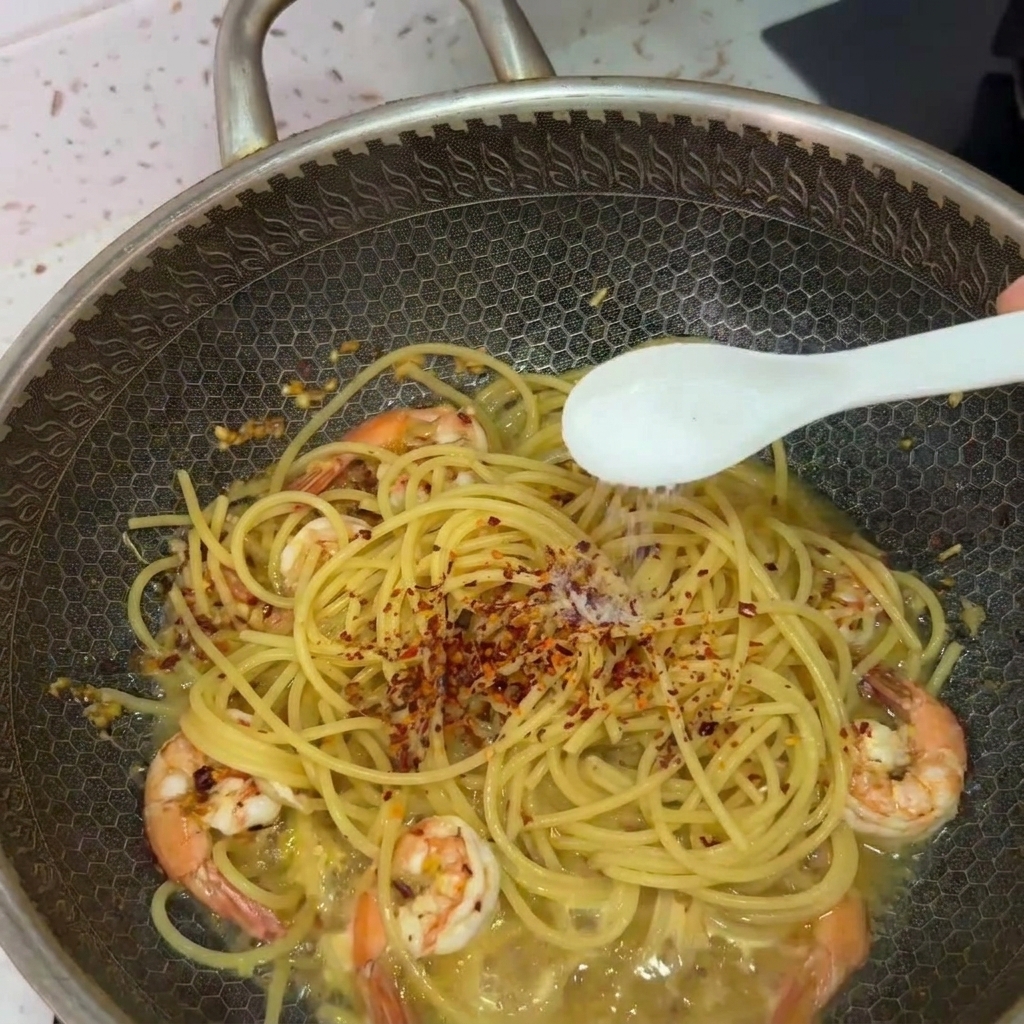 Una cuchara blanca esparciendo sal sobre los espaguetis y camarones en un wok, rematados con copos de chile.