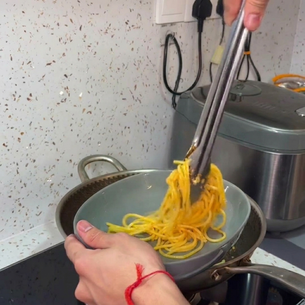 Manos usando pinzas de metal para transferir y girar espaguetis calientes desde una sartén a un bol de servicio gris.