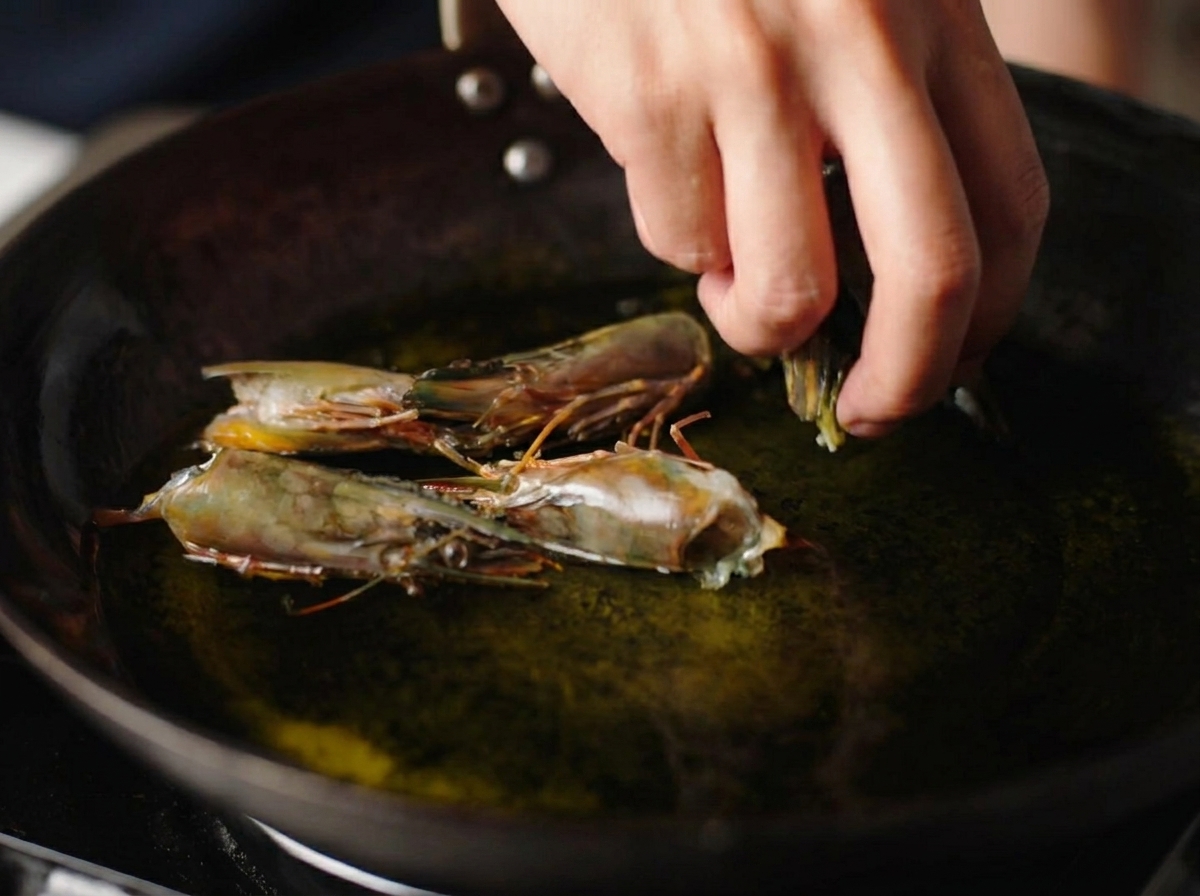 Una mano añadiendo cabezas de gambas crudas en un wok con aceite caliente brillante.