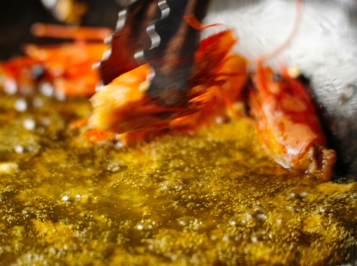 Pinzas levantando una cabeza de gamba frita de una sartén con aceite infusionado de color naranja.