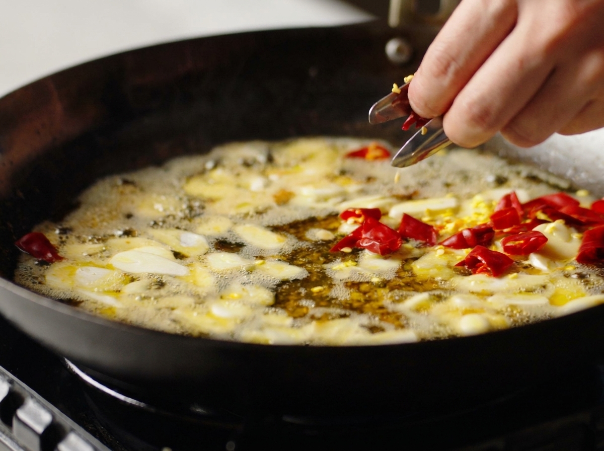 Una mano echando trozos de chile seco en un wok con ajo y aceite sazonado.