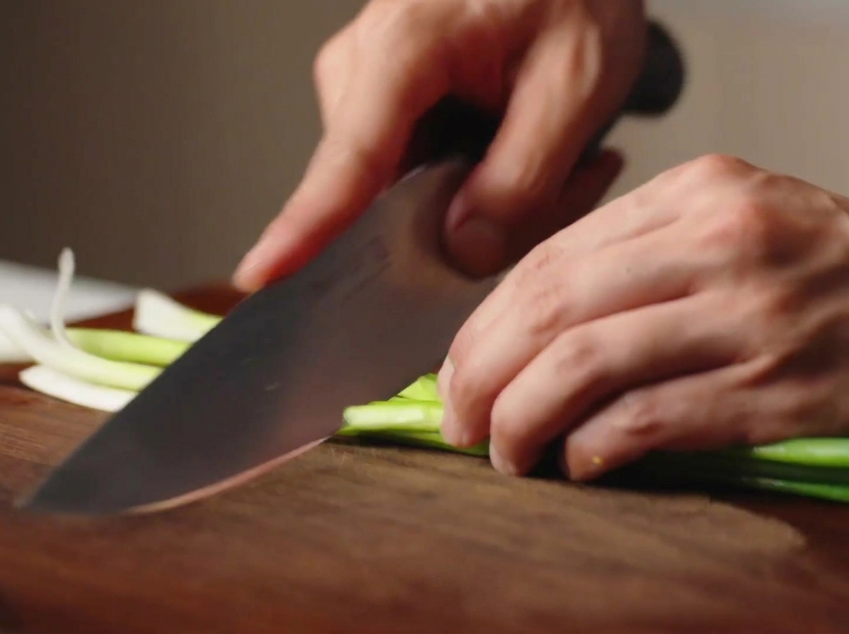 Manos usando un cuchillo de chef para picar cebolletas frescas en trozos sobre una tabla de madera.