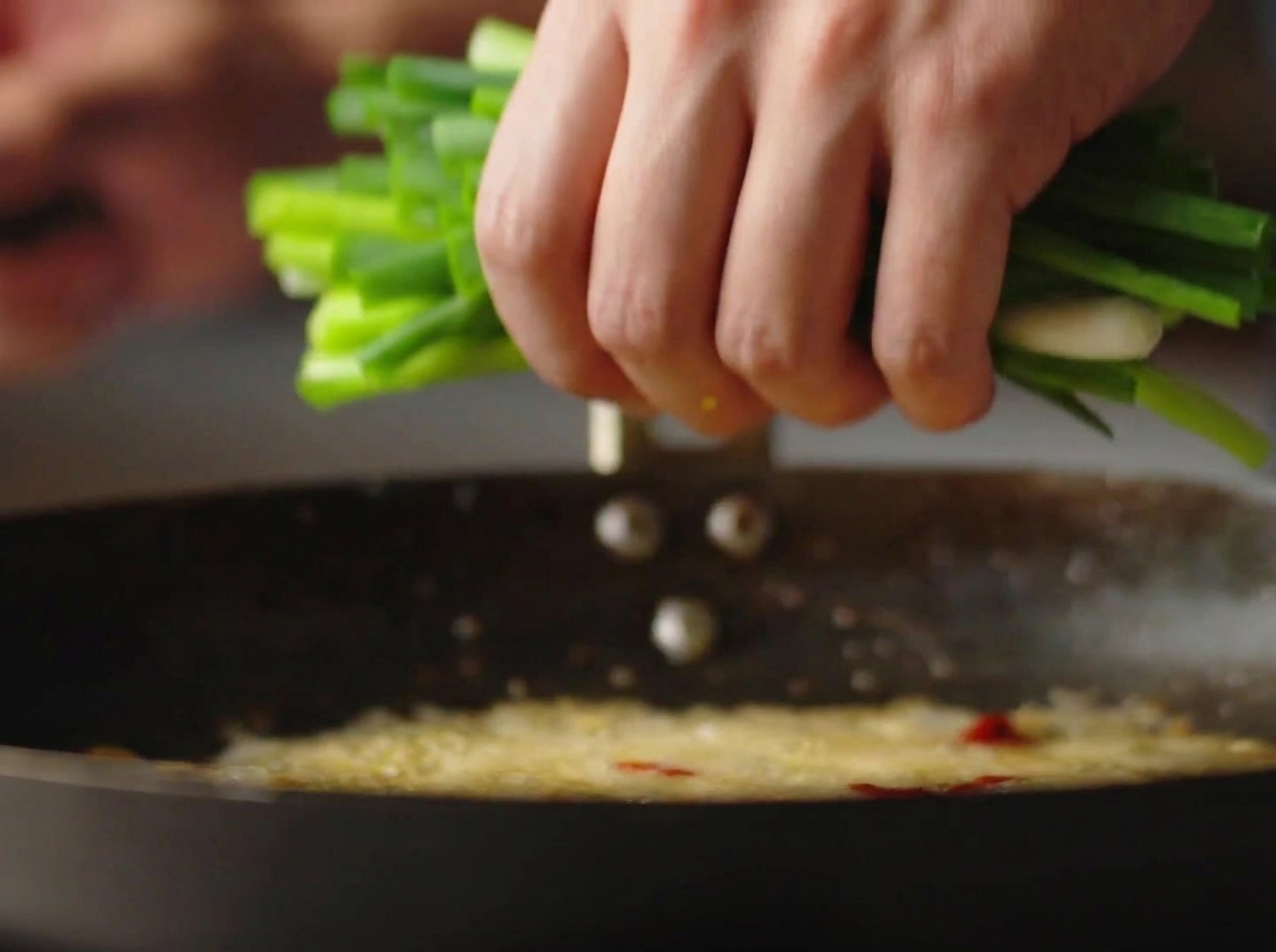 Una mano echando un manojo de cebolleta picada en un wok caliente con aceite burbujeante.