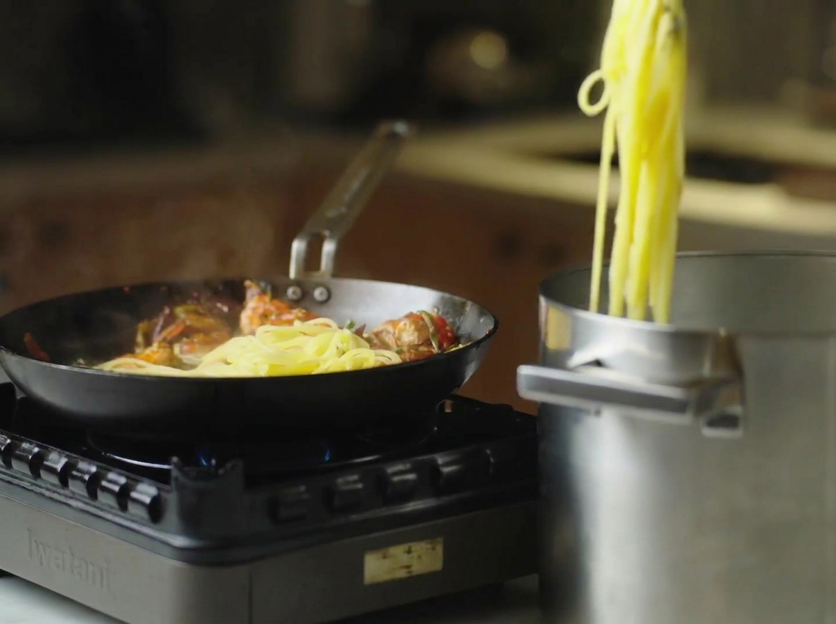 Pinzas levantando espaguetis cocidos de una olla y pasándolos a una sartén con gambas.