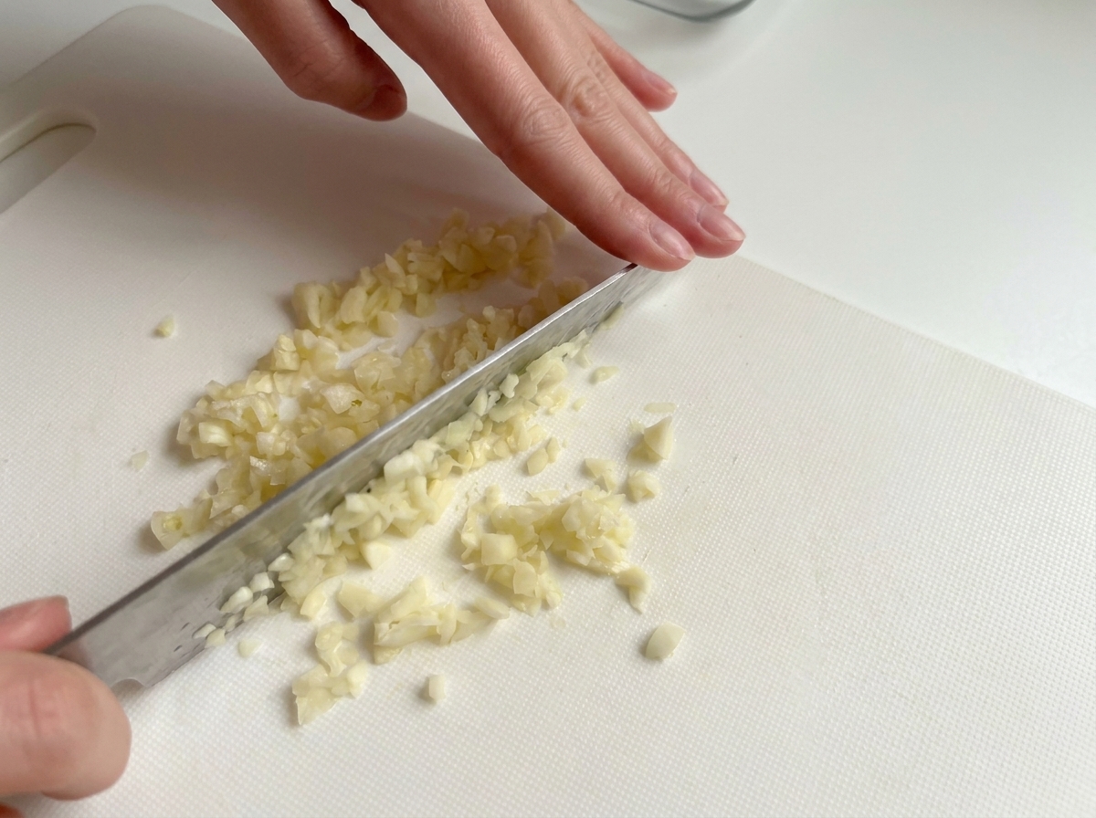 Manos usando un cuchillo para picar finamente dientes de ajo en una tabla de cortar blanca.