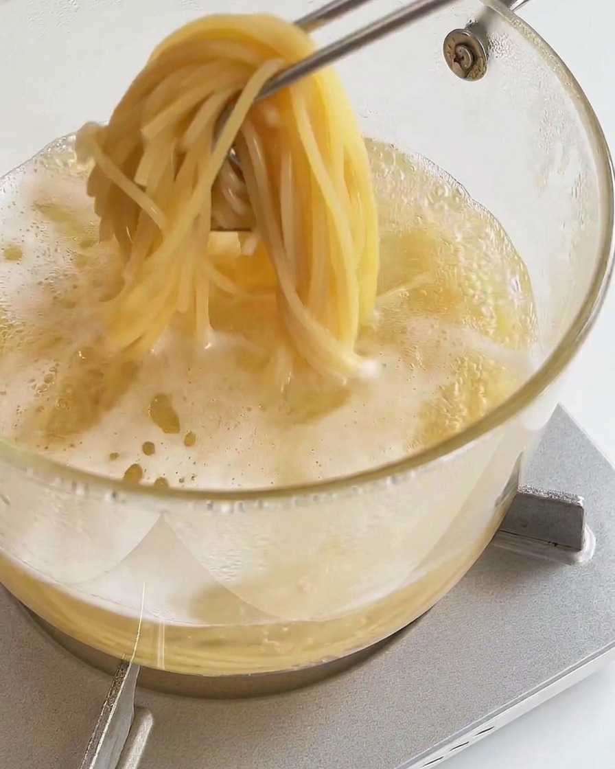 Unas pinzas de cocina sacando una ración de espaguetis cocidos de una olla con agua hirviendo.