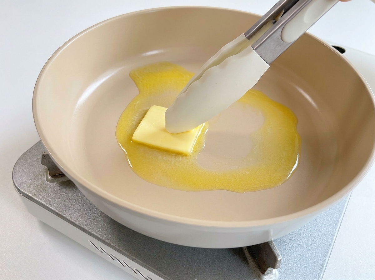 Un dado de mantequilla derritiéndose en una sartén caliente, movido por unas pinzas de silicona.