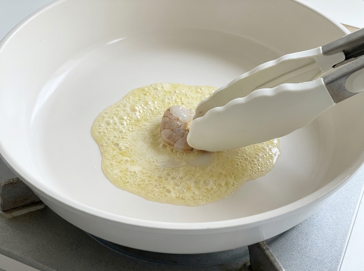 Unas pinzas de silicona colocando un langostino crudo en una sartén con mantequilla derretida burbujeante.