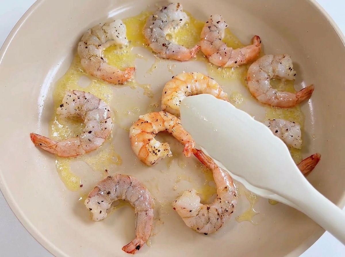 Pinzas blancas levantando un langostino rosado cocido de una sartén con otros langostinos y mantequilla.