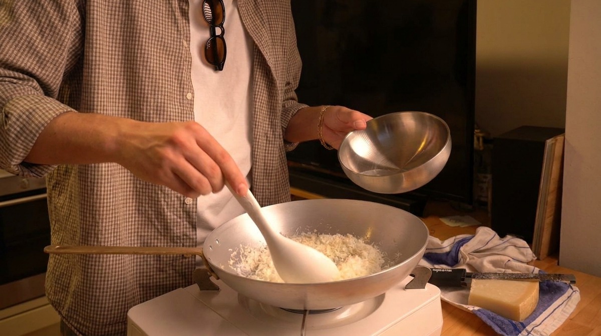 Vertiendo agua templada desde un pequeño bol plateado en una sartén con queso Pecorino recién rallado.
