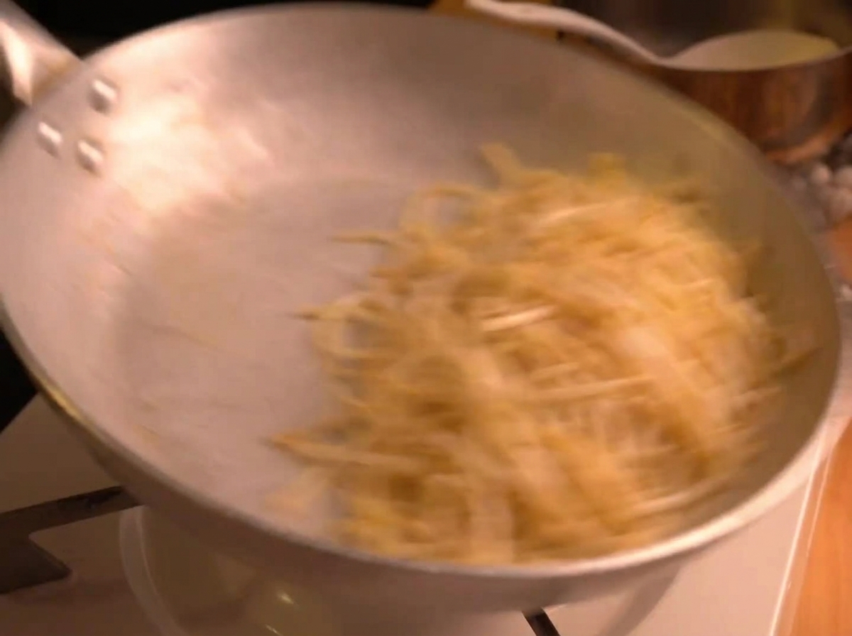 Espaguetis siendo salteados vigorosamente en una sartén plateada, creando un efecto de movimiento mientras la salsa se emulsiona.