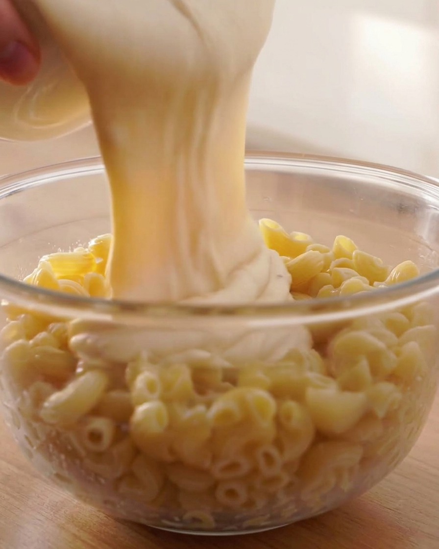 Salsa de queso cremosa vertiéndose sobre macarrones en espiral cocidos en un bol de cristal.