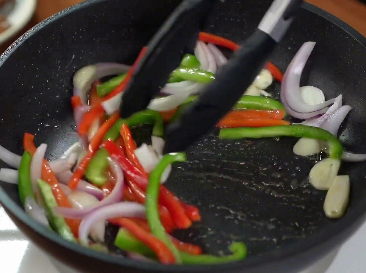 Pinzas de cocina negras salteando cebolla roja y pimientos verdes y rojos en una sartén caliente.