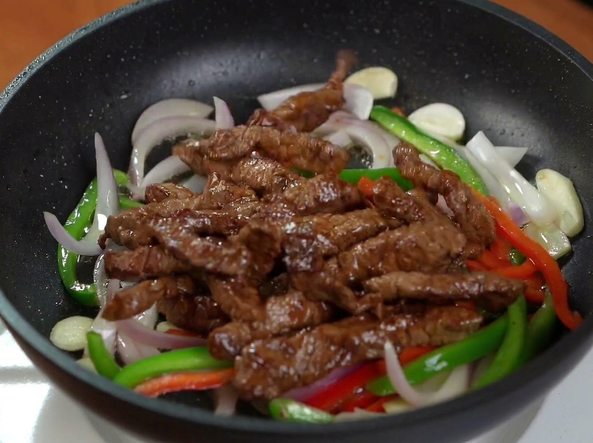 Tiras de carne doradas añadidas a una sartén negra que contiene cebolla roja y pimientos salteados.