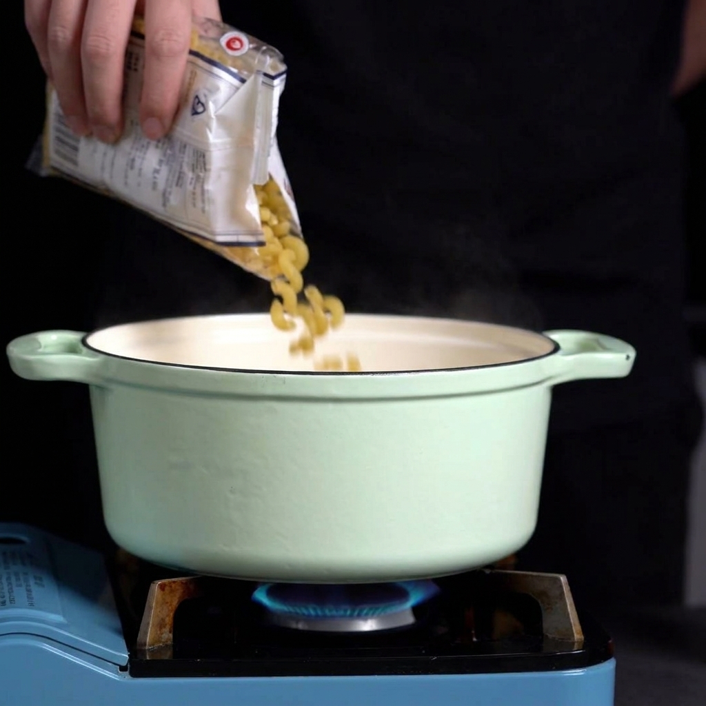 Manos vertiendo pasta de coditos seca desde una bolsa de plástico transparente en una olla verde con agua hirviendo.