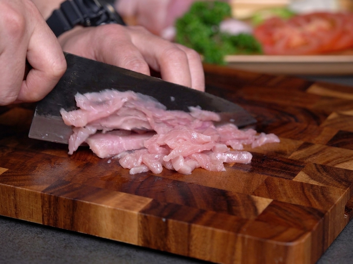 Manos cortando carne magra rosada en tiras finas sobre una tabla de madera con un cuchillo oscuro.