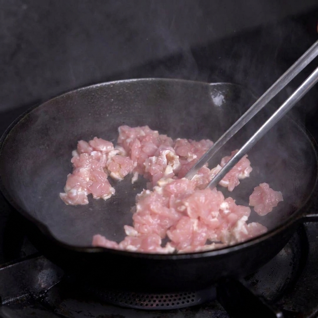 Carne magra en dados cocinándose en una sartén de hierro caliente con pinzas metálicas.