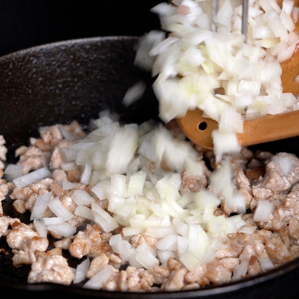 Cebolla blanca picada cayendo desde una tabla a una sartén con carne parcialmente cocinada.
