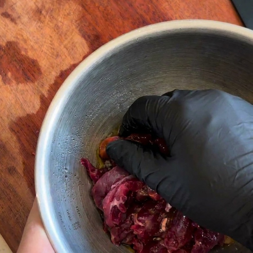 Mano con guante mezclando minuciosamente las tiras de ternera con el marinado oscuro en un bol.