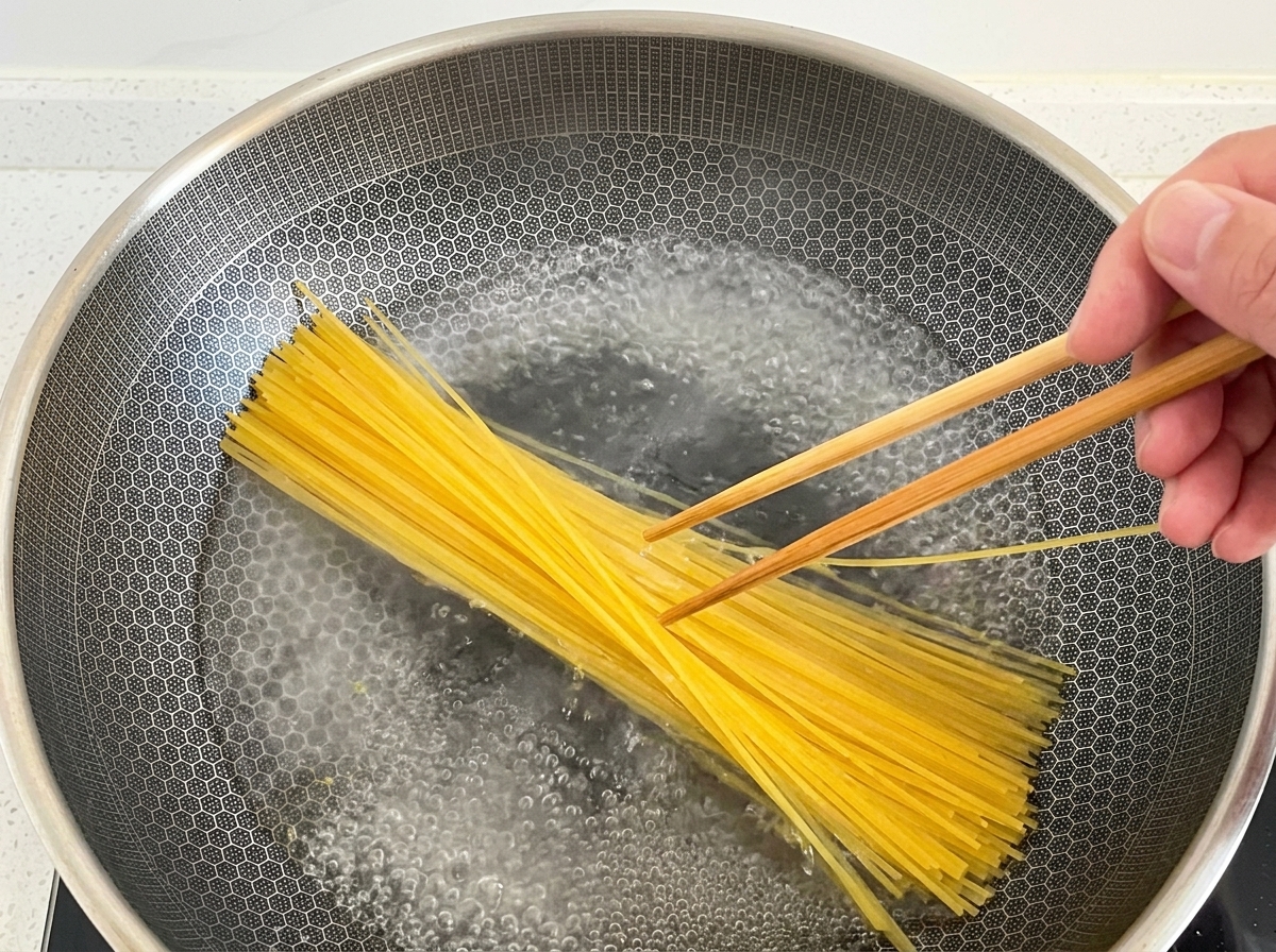 Mano usando palillos de madera para sumergir espaguetis secos en un wok grande con agua hirviendo.