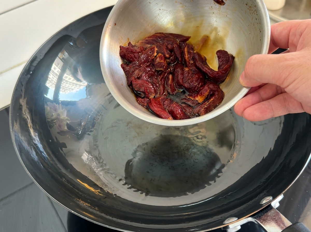 Tiras de carne marinada cayendo desde un bol metálico en un wok caliente con aceite.
