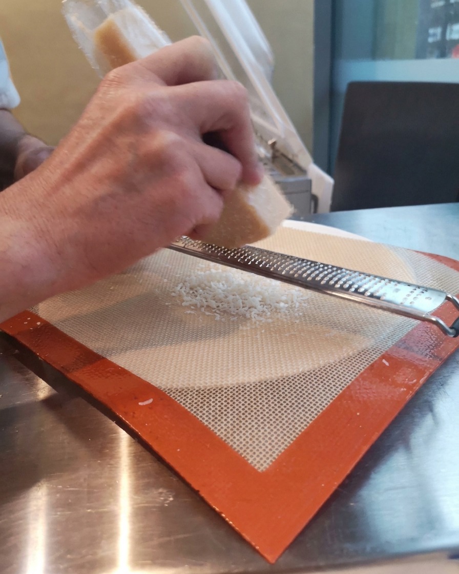 Manos rallando un bloque de queso parmesano sobre un tapete de silicona usando un rallador tipo microplane largo.