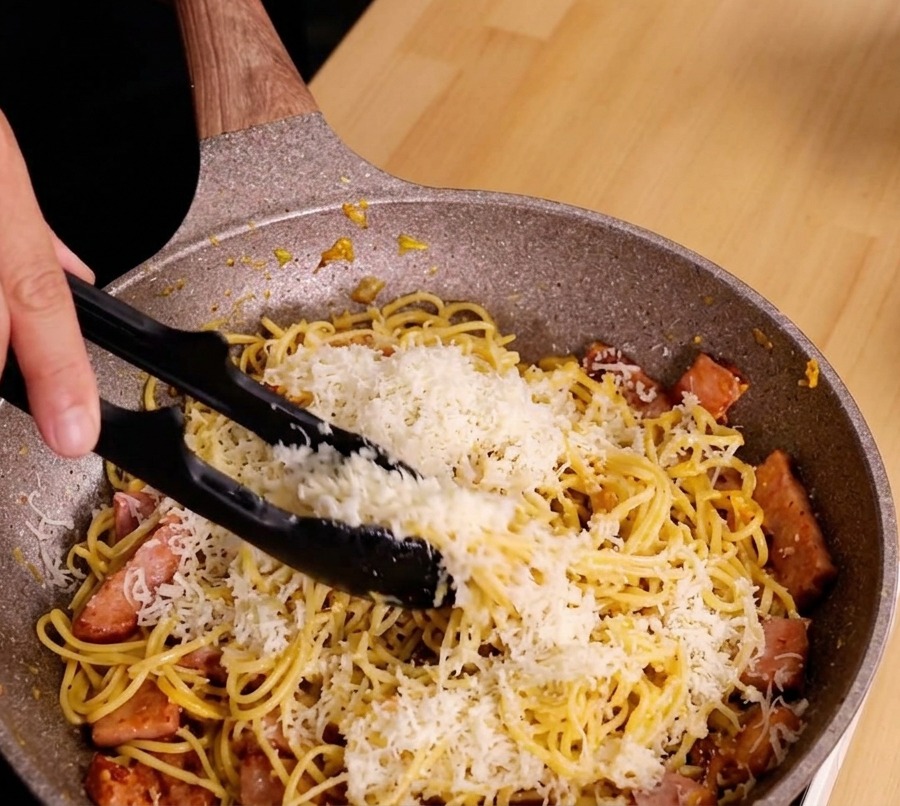 Manos usando pinzas para remover vigorosamente los espaguetis, el queso y el bacon en una sartén moteada para crear una salsa cremosa.