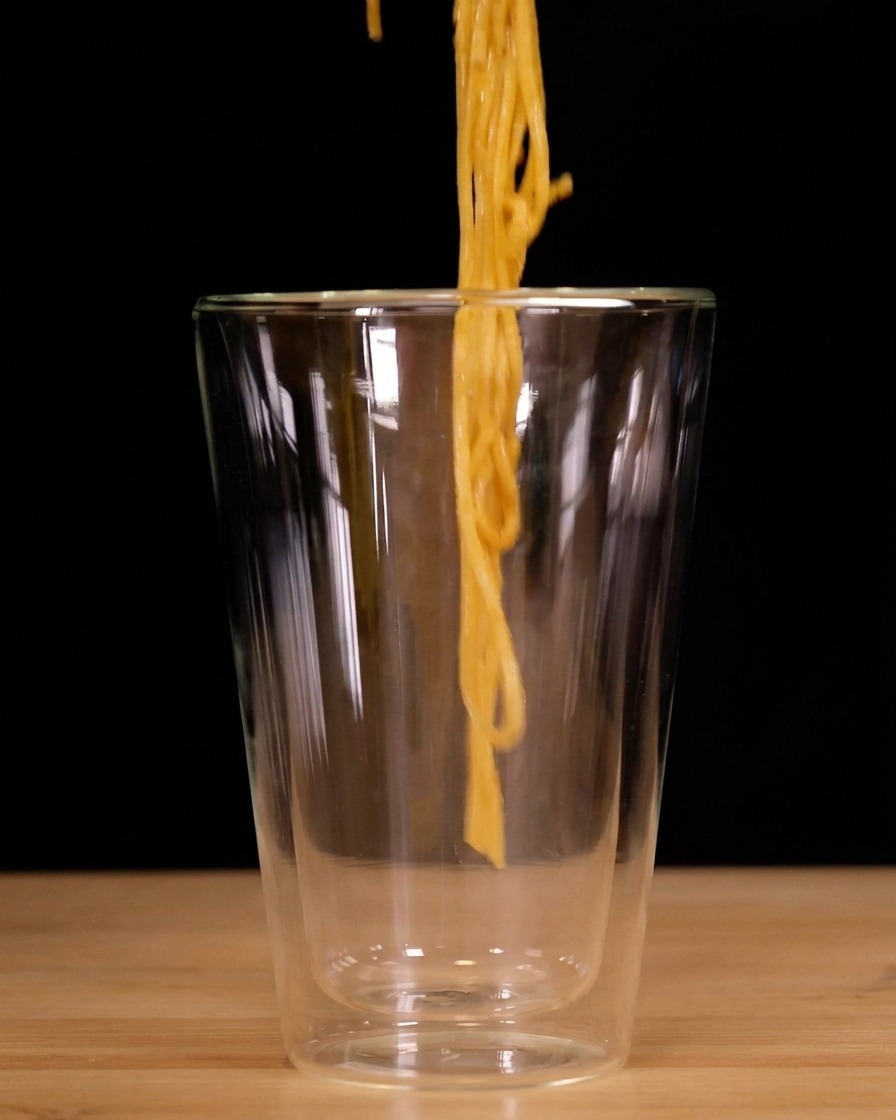 Espaguetis enrollados verticalmente con pinzas y bajados a un vaso alto de doble pared transparente.