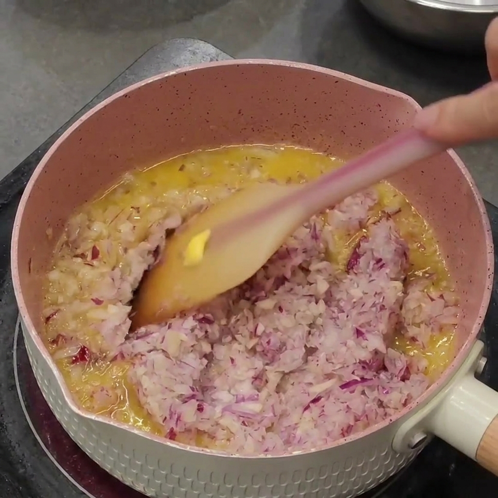 Cebolla morada y ajo picados siendo sofreídos en mantequilla derretida dentro de una cacerola rosa.