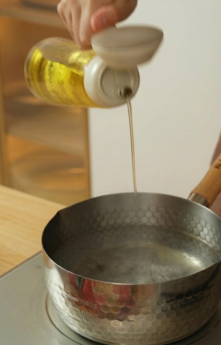 Vertiendo aceite de cocina desde una botella en una olla de acero inoxidable con agua caliente.