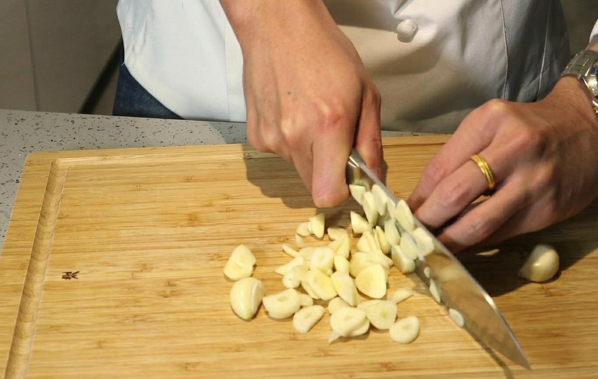 Manos de un chef usando un cuchillo para picar toscamente dientes de ajo sobre una tabla de madera.