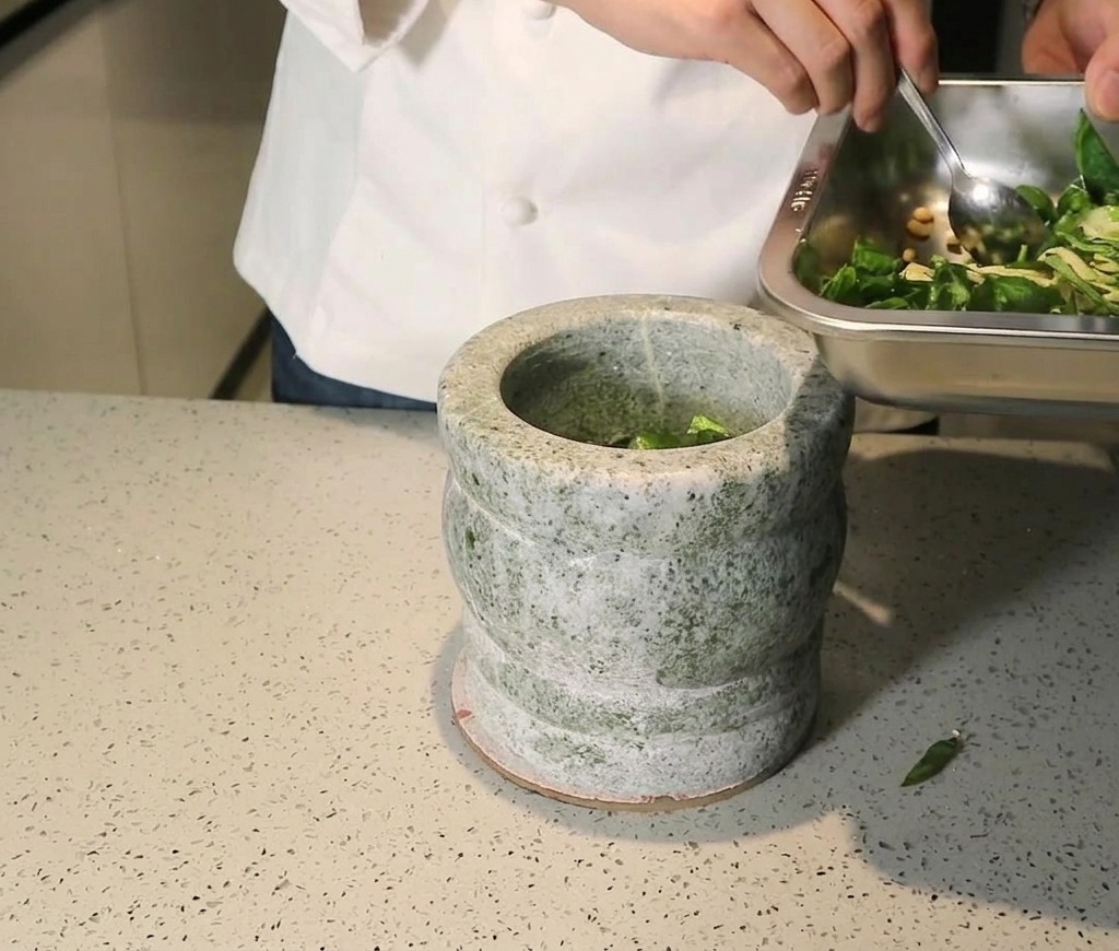 Chef pasando una mezcla de albahaca fresca, piñones y ajo desde una bandeja metálica a un mortero de piedra gris.