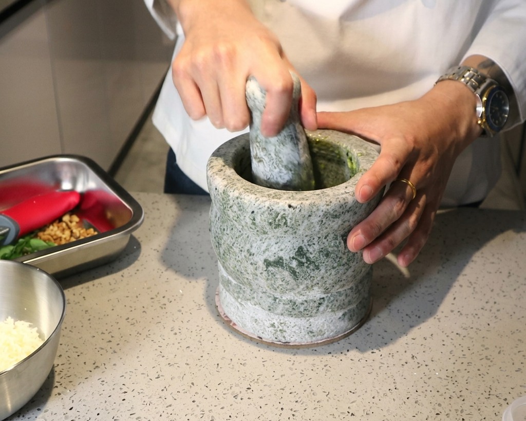 Una persona usando un mortero y mazo de piedra para triturar ingredientes frescos hasta formar una pasta de pesto verde.