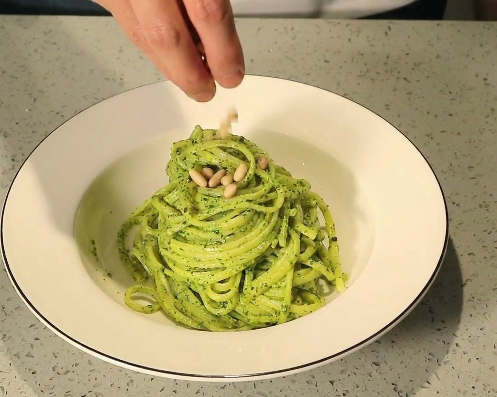Dedos espolvoreando piñones tostados sobre un nido de pasta al pesto verde en un bol de cerámica blanca.