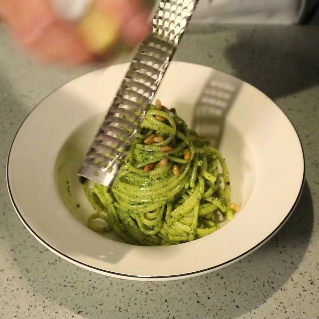 Una mano usando un rallador metálico para sacar lascas de queso parmesano fresco sobre un bol de pasta al pesto con piñones.