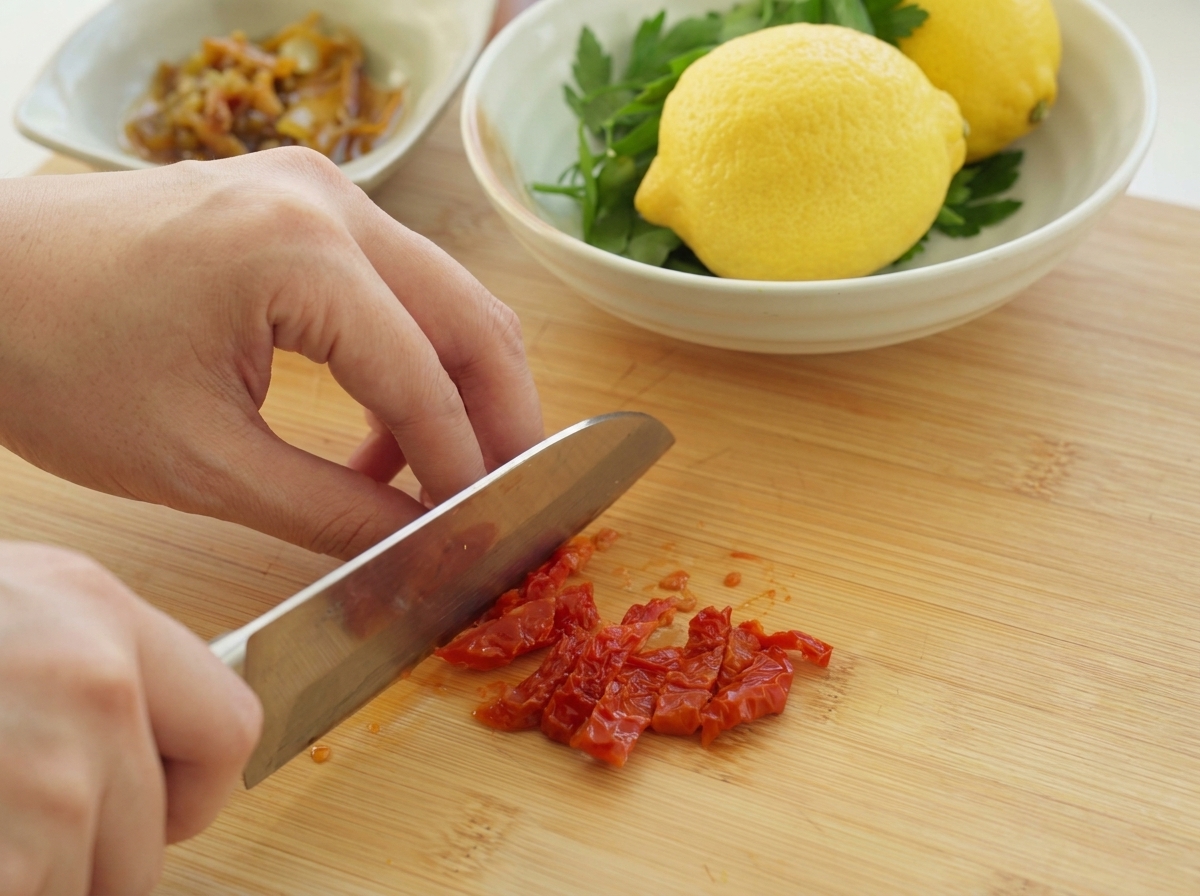 Una persona usando un cuchillo de chef para picar finamente tomates secos rojos sobre una tabla de madera.