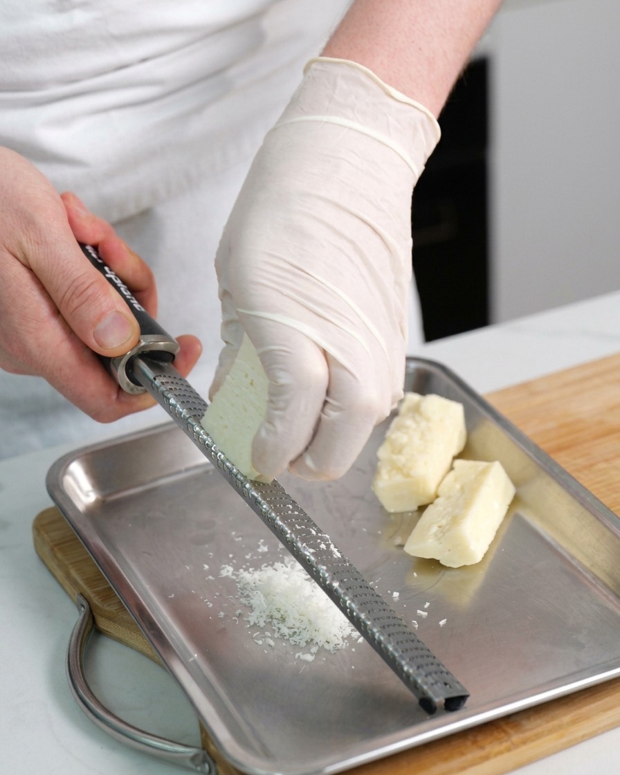 Manos rallando un bloque de queso Pecorino Romano sobre una bandeja metálica.
