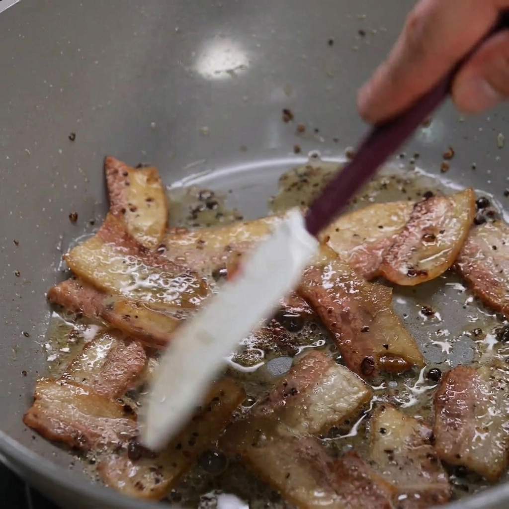 Tiras de guanciale dorándose en una sartén mientras sueltan su grasa.