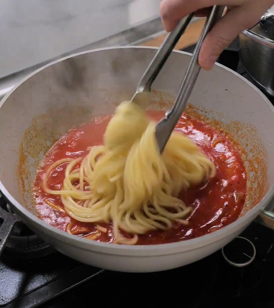 Pinzas metálicas pasando bucatini cocidos directamente a la sartén con salsa de tomate.