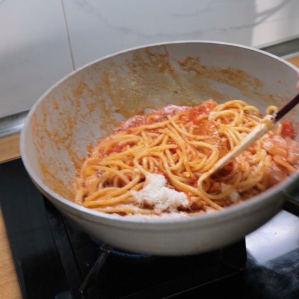 Mezclando bucatini con salsa de tomate roja y queso fundido en una sartén blanca.