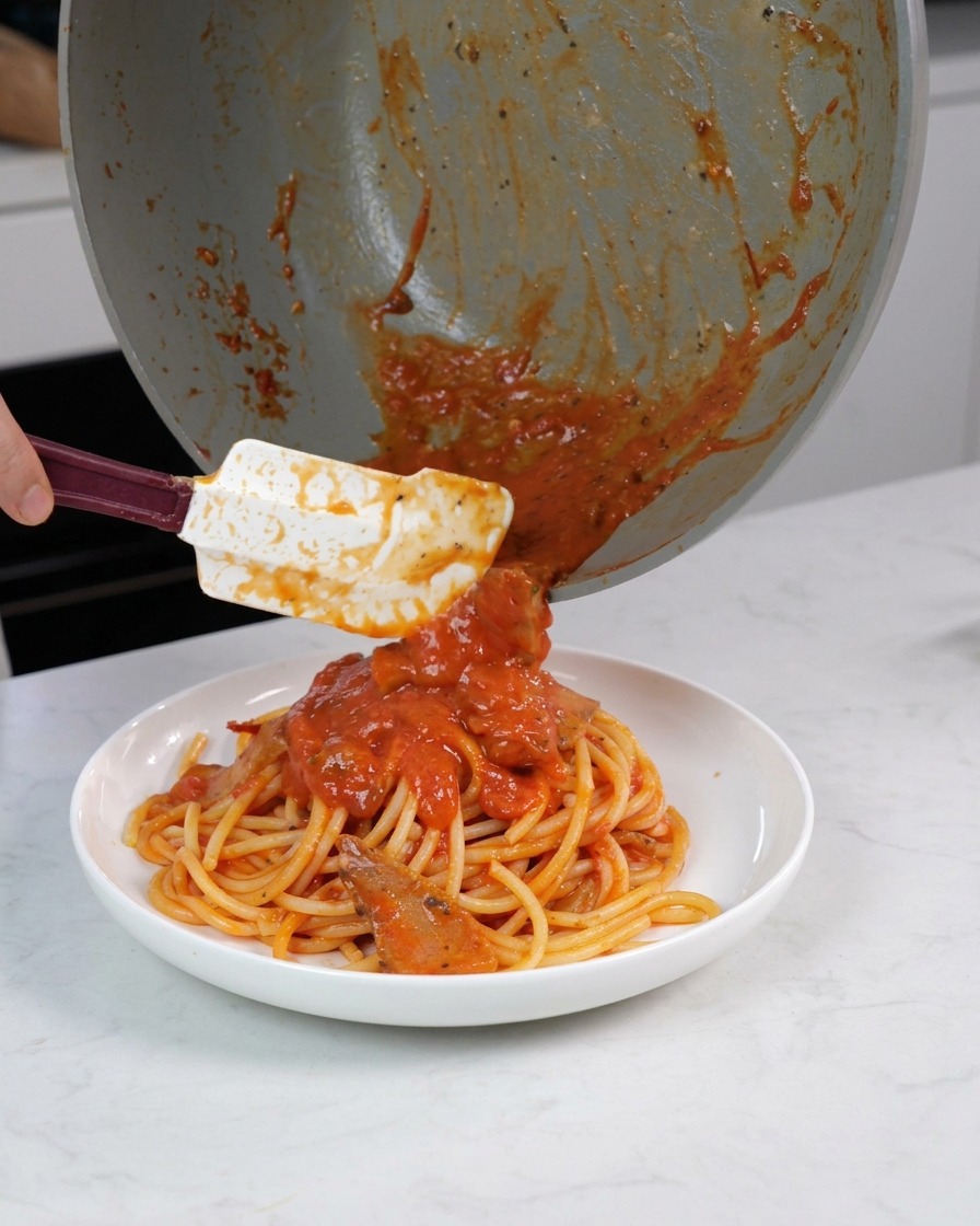 Usando una espátula para servir el resto de la salsa con trozos de guanciale sobre la pasta emplatada.