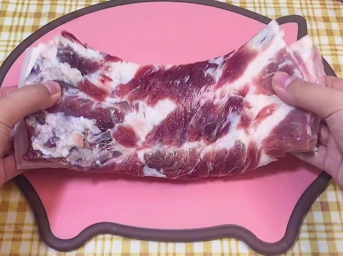 Manos sosteniendo una pieza de panceta cruda sobre una tabla de cortar rosa con forma de cerdo.