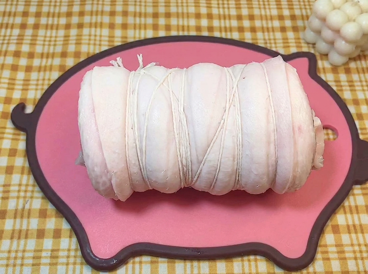Cilindro de panceta cruda atado firmemente con hilo de cocina sobre una tabla rosa.